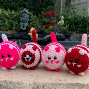May include: Four crocheted cat toys in pink, red, and white with striped patterns. The cats have black eyes and pink noses.