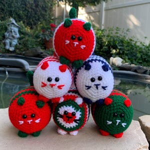 May include: A stack of six crocheted cat toys in various colors, including red, white, green, and blue. Each toy has a cat face with black eyes and a white nose.