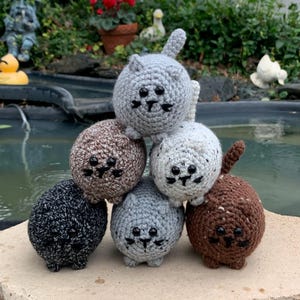 May include: A stack of six crocheted cat toys in various colors: gray, brown, black, and white. Each cat has a round body, small ears, and a stitched face with black eyes and whiskers. The toys are arranged on a stone surface.