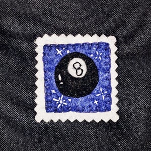 May include: A handmade felt patch in the shape of a postage stamp. The patch features a black eight ball on a blue background with white star accents. The edges are white with a scalloped design.