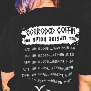 May include: Black t-shirt with white text that reads "Corroded Coffin 1986 NMOO 3015AN TOUR" and a list of tour dates and locations. The text is in a grunge style font.