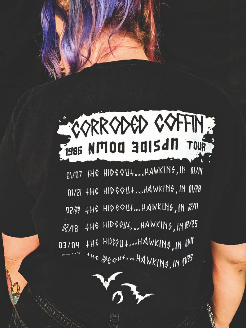 May include: Black t-shirt with white text that reads "Corroded Coffin 1986 NMOO 3015AN TOUR" and a list of tour dates and locations. The text is in a grunge style font.
