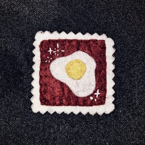 May include: A square, felt patch with a fried egg design. The egg is white with a yellow yolk, set against a burgundy background. The patch has a white, scalloped border and small white starburst accents.
