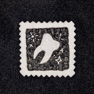 May include: A white felt patch with a black embroidered square featuring a white tooth and starburst designs. The patch has a white, scalloped border and is set against a dark, textured background.