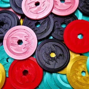 May include: A collection of colorful round tokens with various designs. The tokens are made of plastic and have a raised design. The colors include red, black, gold, pink, and teal.