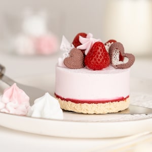 May include: A small pink cake with a strawberry, chocolate hearts, and a pink swirl on top. The cake is on a white plate with two small meringue cookies.