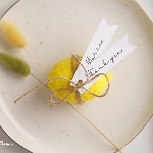 May include: A yellow felt lemon with a brown twine bow and two white tags. The tags are tied to the bow and have the text "Maria" and "Thank you" written on them.