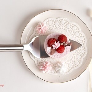 May include: A small pink cake with strawberries and a white meringue on top, on a white plate with a lace doily, and a silver cake server.