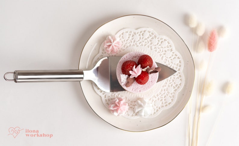 May include: A small pink cake with strawberries and a white meringue on top, on a white plate with a lace doily, and a silver cake server.