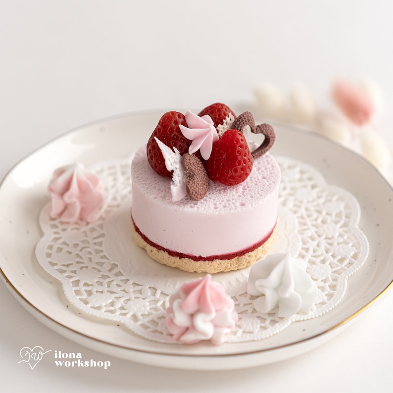 May include: A small pink cake with a white frosting and a strawberry and chocolate topping. The cake is on a white plate with a doily and pink and white meringue cookies.