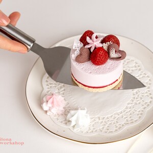 May include: A small pink cake with white frosting and red strawberries on top. The cake is on a white plate with a white doily. A silver cake server is holding the cake.