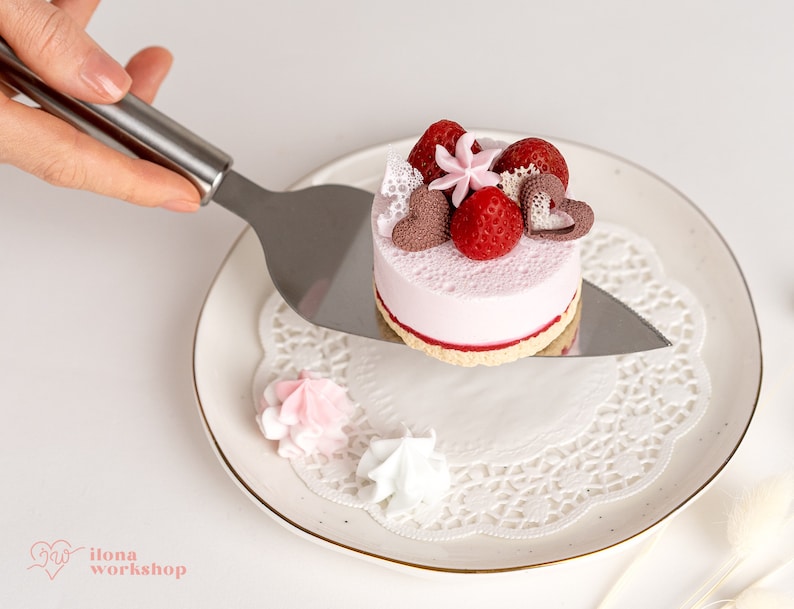 May include: A small pink cake with white frosting and red strawberries on top. The cake is on a white plate with a white doily. A silver cake server is holding the cake.