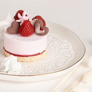 May include: A small pink cake with a strawberry and chocolate heart topping, on a white plate with a lace doily. The cake is surrounded by white and pink meringue.