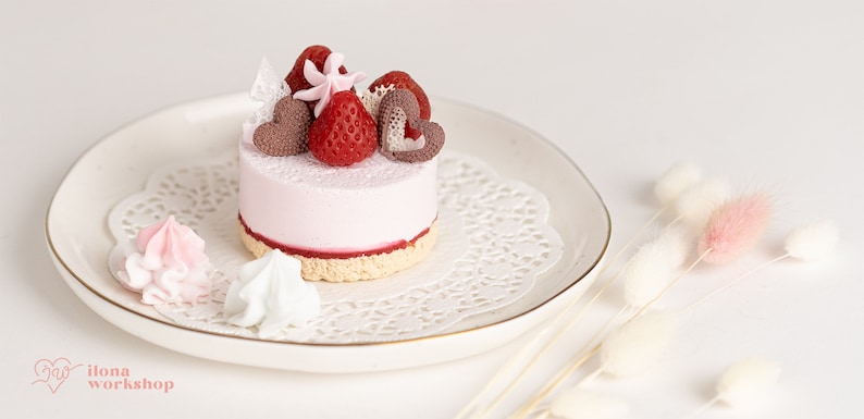 May include: A small pink cake with a strawberry and chocolate heart topping, on a white plate with a lace doily. The cake is surrounded by white and pink meringue.