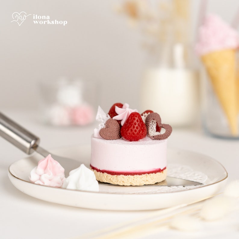 May include: A small pink cake with a strawberry, chocolate hearts, and a pink swirl on top. The cake is on a white plate with two small meringue cookies.