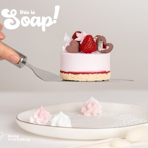 May include: A pink and white cake-shaped soap with a strawberry and chocolate topping. The soap is on a silver spatula and the text "This is Soap!" is in the background.