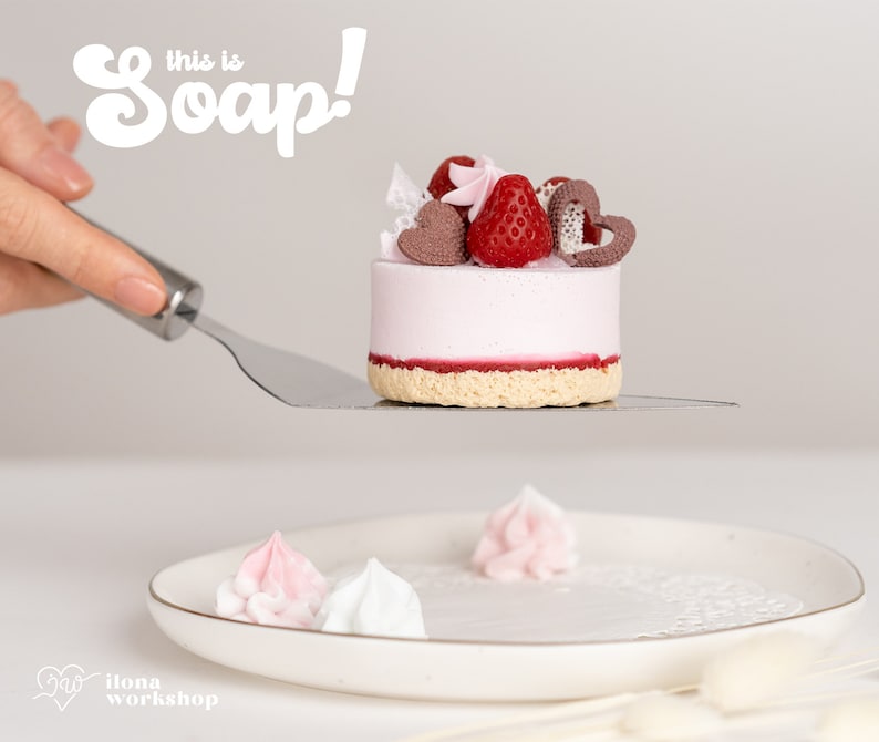 May include: A pink and white cake-shaped soap with a strawberry and chocolate topping. The soap is on a silver spatula and the text "This is Soap!" is in the background.