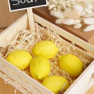 May include: A wooden crate filled with yellow lemon-shaped soaps. The crate is lined with shredded paper and has a small chalkboard sign that says "Soap".