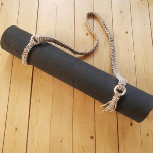May include: A rolled black yoga mat held together by a beige macrame strap. The strap has wooden rings and a woven pattern. The mat is on a light wooden floor, creating a neutral and natural aesthetic.