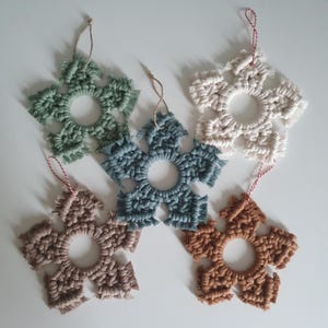 Könnte beinhalten: Fünf handgefertigte Makramee-Schneeflocken-Ornamente in verschiedenen Farben, darunter Grün, Blau, Weiß und Braun. Jede Schneeflocke hat eine kreisförmige Öffnung in der Mitte und ist mit einer Schnur zum Aufhängen versehen.