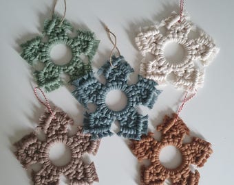 Könnte beinhalten: Fünf handgefertigte Makramee-Schneeflocken-Ornamente in verschiedenen Farben, darunter Grün, Blau, Weiß und Braun. Jede Schneeflocke hat eine kreisförmige Öffnung in der Mitte und ist mit einer Schnur zum Aufhängen versehen.