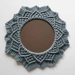 May include: A round mirror with a blue macrame frame. The frame is made of knotted cords and has a fringe around the edge. The mirror is ideal for adding a bohemian touch to any room.