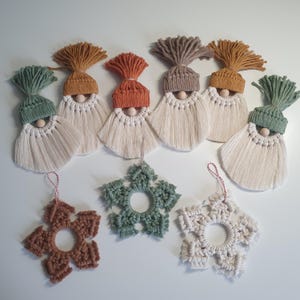 Op de afbeelding: Een verzameling handgemaakte macramé kerstversieringen. De ornamenten omvatten kabouterfiguren met kleurrijke hoeden en baarden, en stervormige sneeuwvlokken in verschillende kleuren. De kabouters hebben houten kralen neuzen en de sneeuwvlokken hebben ophangtouwtjes.