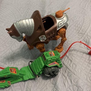 May include: A brown and silver toy horse with a saddle and a green vehicle with orange accents. The toy horse has articulated legs and a detailed head. The green vehicle has a large wheel and a red pull cord.