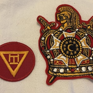 May include: Two embroidered patches. One is a red circle with a gold triangle and a symbol inside. The other is a red patch in the shape of a crown with gold and black embroidery, a starburst design, and swords.