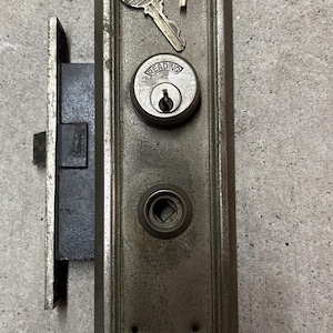 May include: A vintage brass door lock with a key and a keyhole. The lock has a rectangular plate with a rounded edge and a small square hole in the center. The lock is labeled "READING".
