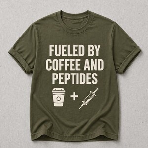May include: An olive green short-sleeve t-shirt featuring the cream-colored text FUELED BY COFFEE AND PEPTIDES. Below the text is a graphic of a coffee cup, a plus sign, and a syringe. The shirt has a crew neckline and a standard fit design.