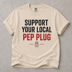 May include: A beige short-sleeve t-shirt with the black text SUPPORT YOUR LOCAL and the red text PEP PLUG printed on the front. Below the text is a small black icon of a medicine vial with a red plus sign, flanked by two horizontal lines.