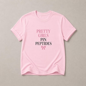 May include: A light pink short-sleeved t-shirt featuring the text PRETTY GIRLS in pink, followed by PIN PEPTIDES in black, and a small pink bow icon centered below the text. The shirt has a crew neckline and cuffed sleeves.