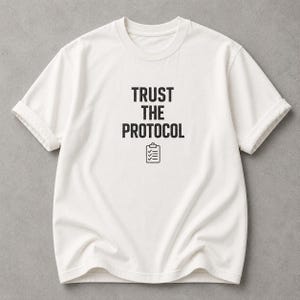 May include: A short-sleeved, off-white crew neck t-shirt featuring the black text "TRUST THE PROTOCOL" printed on the center of the chest, positioned above a simple black line-art icon of a clipboard with three checkmarks.