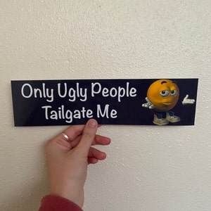 May include: A black and blue bumper sticker with white text that reads "Only Ugly People Tailgate Me". A yellow cartoon face with a frown and a raised eyebrow is on the right side of the sticker.