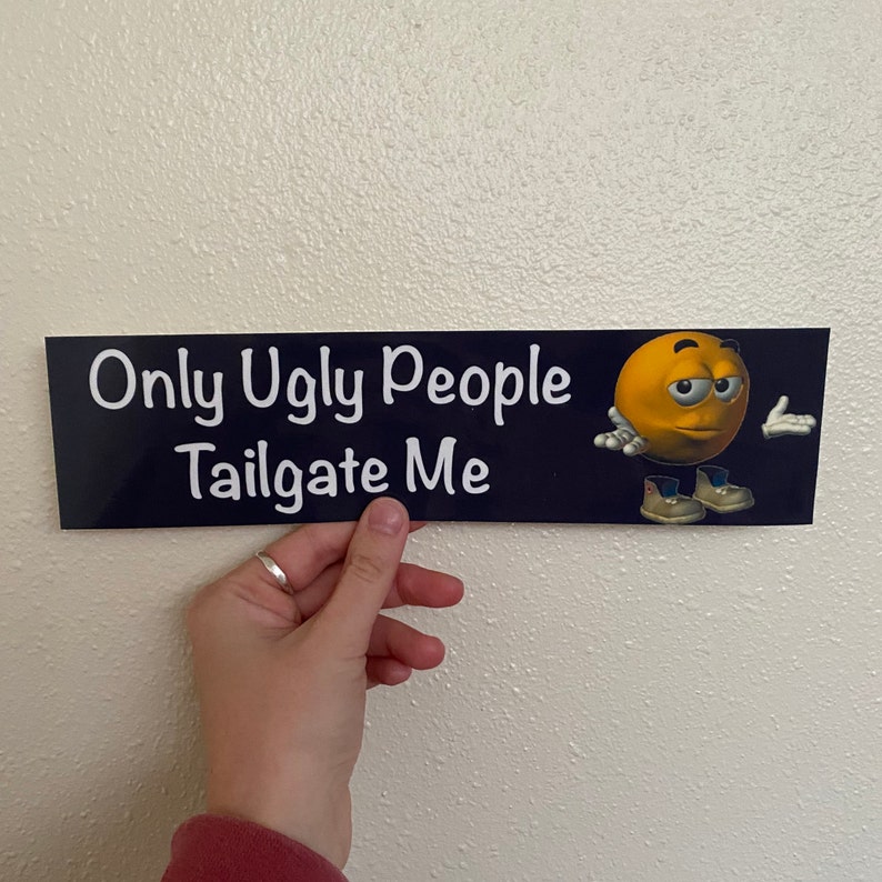 May include: A black and blue bumper sticker with white text that reads "Only Ugly People Tailgate Me". A yellow cartoon face with a frown and a raised eyebrow is on the right side of the sticker.