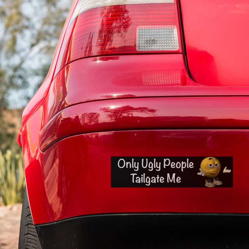 May include: A red car with a bumper sticker that says "Only Ugly People Tailgate Me" with a yellow cartoon face with a frown and a hand gesture.