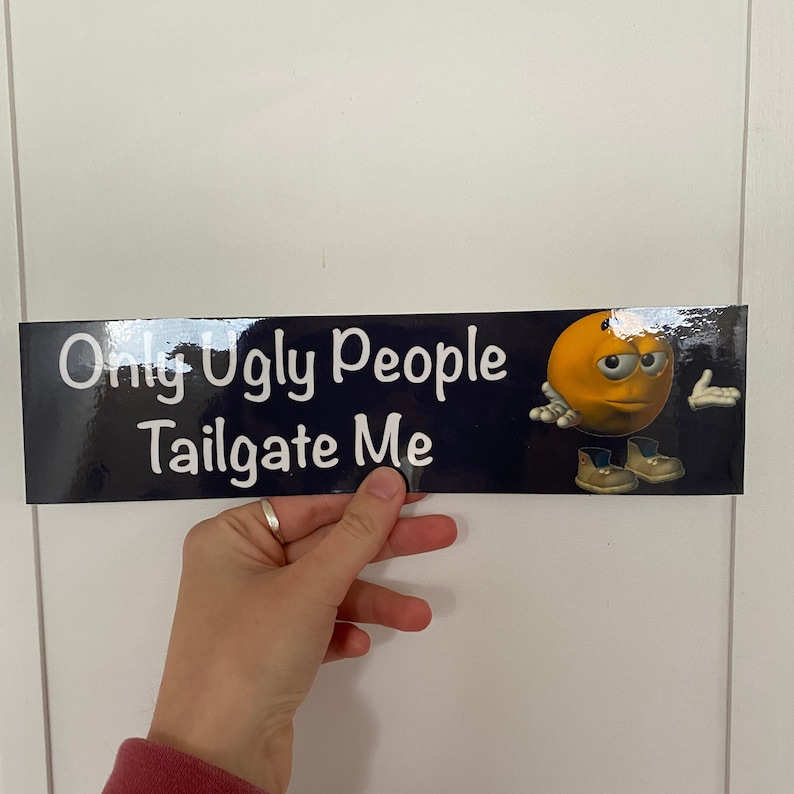 May include: A black and blue bumper sticker with white text that reads "Only Ugly People Tailgate Me". A yellow cartoon face with a frown is on the right side of the sticker.