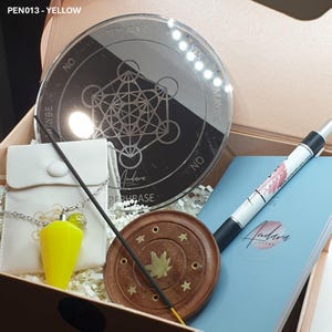 May include: A gift box set with a yellow pendulum, a wooden incense burner, a blue notebook, a pen, and a silver mirror with a geometric design and the words "Rephrase" and "Yes/Maybe/No".