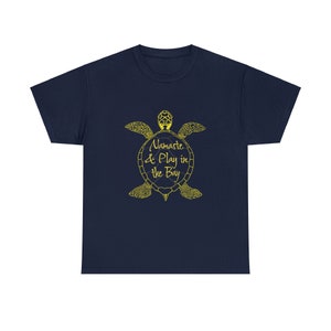 Puede incluir: Camiseta azul marino con un contorno amarillo de una tortuga marina y el texto "Namaste & Play in the Bay".