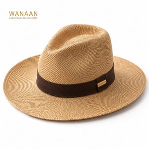 Op de afbeelding: Een beige fedora hoed van gevlochten stro met een brede rand en een donkerbruine stoffen band rond de kroon. Een klein rechthoekig goudkleurig metalen plaatje met de tekst WANAAN is bevestigd aan de zijkant van de bruine band op dit hoofddeksel.