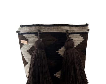 Puede incluir: Un bolso tejido con un patrón geométrico en tonos marrones y grises. El bolso presenta dos grandes borlas marrón oscuro y una pequeña etiqueta con texto. El bolso está diseñado con una forma redondeada y una construcción robusta.