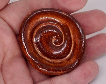 May include: A small, round, ceramic dish with a brown glaze and a spiral design. The dish is glazed in a light brown color with a darker brown spiral design.
