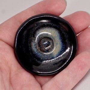 Puede incluir: Un plato de cerámica negro con un esmalte azul y blanco. El plato tiene una forma circular y un borde ligeramente elevado.
