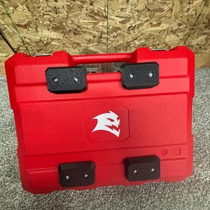May include: A rectangular red plastic storage case featuring a white graphic logo of a stylised creature head on the front. The case includes four black rectangular feet attached with silver screws and a red handle integrated into the top edge.