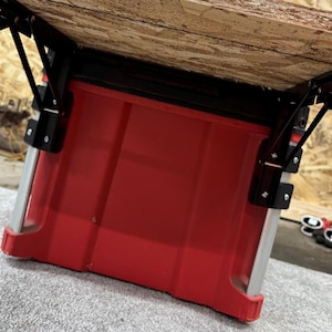 May include: A red plastic storage bin with a black metal frame and a wooden top. The bin is supported by black metal legs and is designed for storage. The image is taken from a low angle, looking up at the underside of the bin.