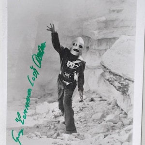 Puede incluir: Fotografía en blanco y negro de una persona con un disfraz de robot, con casco y peto. La persona está de pie en un entorno rocoso al aire libre, con el brazo levantado. La imagen está firmada con tinta verde. El disfraz tiene un cinturón con una hebilla decorativa.