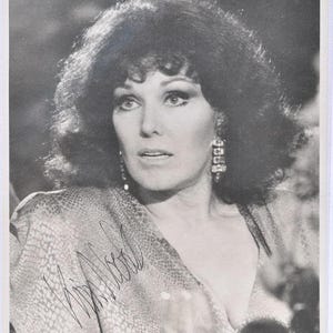 May include: Black and white photograph of a woman with dark, voluminous hair and dramatic eye makeup. She wears a patterned top and large, ornate earrings. The image includes an autograph in the lower left corner.