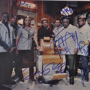 May include: A group of people, likely actors, pose in a barbershop setting. The image features a vintage barber chair, and the subjects are dressed in various outfits. The photo is signed with blue ink.