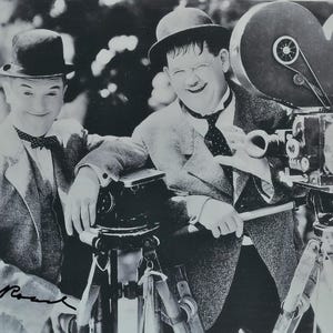 May include: A monochrome photograph featuring Laurel and Hardy, the comedy duo. They are smiling next to a vintage film camera. Both are wearing suits and bowler hats. The image is signed with a handwritten signature.